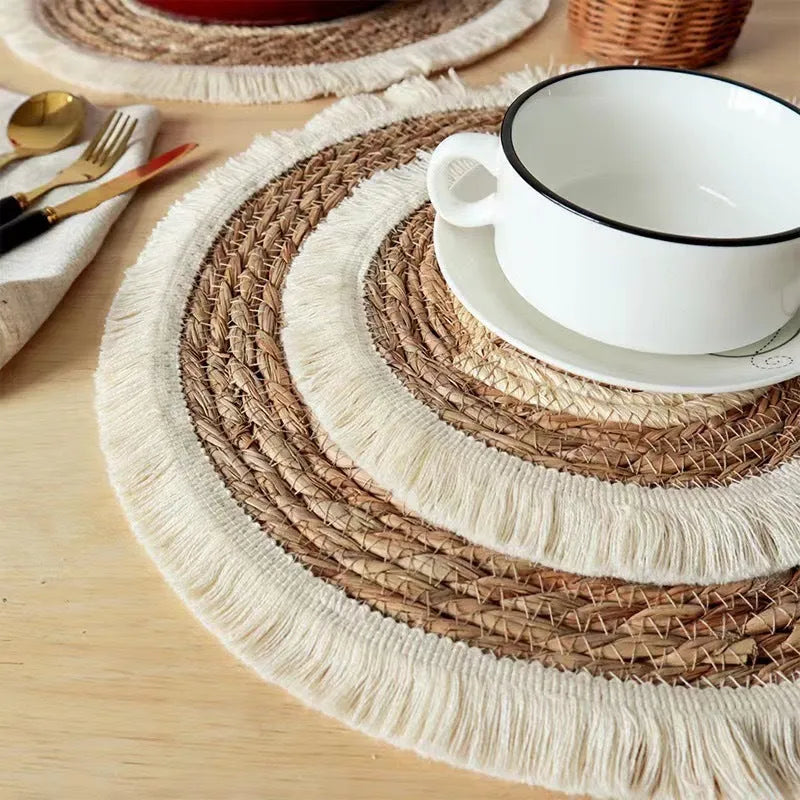 A round natural papyrus placemat with decorative tassels, shown on a wooden table.