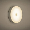 Small round white LED night light with motion sensor, shown with USB charging cable and adhesive tape.