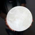 3D printed white moon lamp with detailed craters on a wooden stand, glowing with warm yellow light.