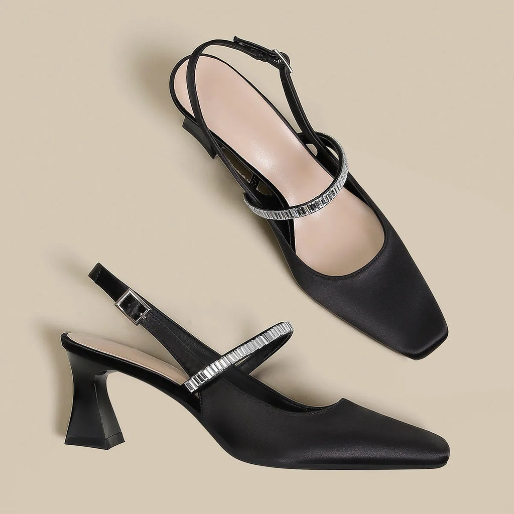 Women's black stiletto heel sandals with pointed closed toe, buckle strap, and skirt detail on a white background