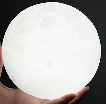 3D printed white moon lamp with detailed craters on a wooden stand, glowing with warm yellow light.