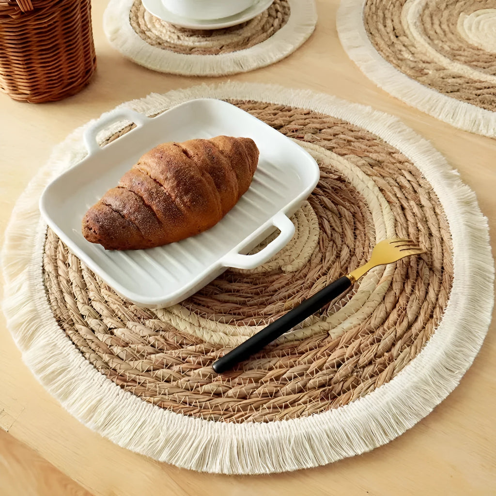 A round natural papyrus placemat with decorative tassels, shown on a wooden table.