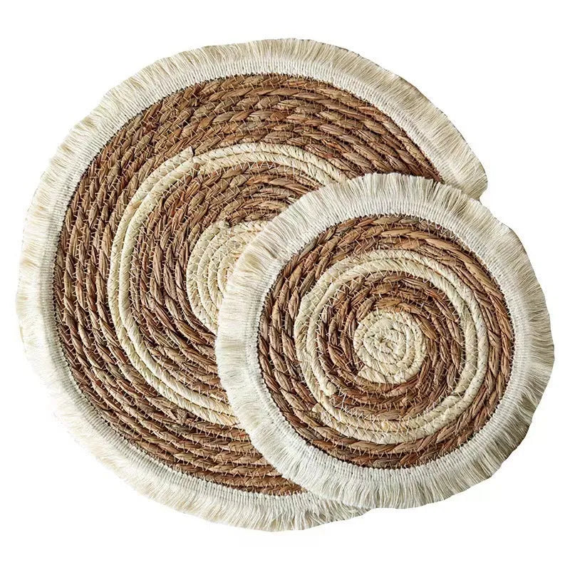 A round natural papyrus placemat with decorative tassels, shown on a wooden table.
